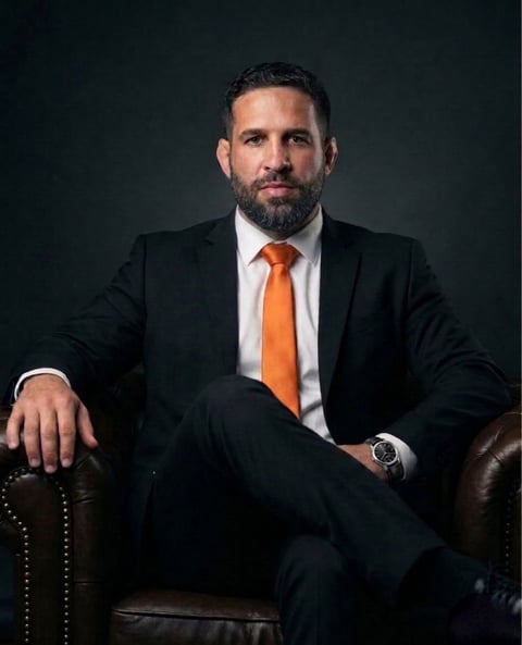 Professional man in dark suit and orange tie sitting on leather chair against dark background, looking at camera