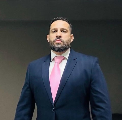 Professional man with beard wearing dark blue suit and pink tie against dark background